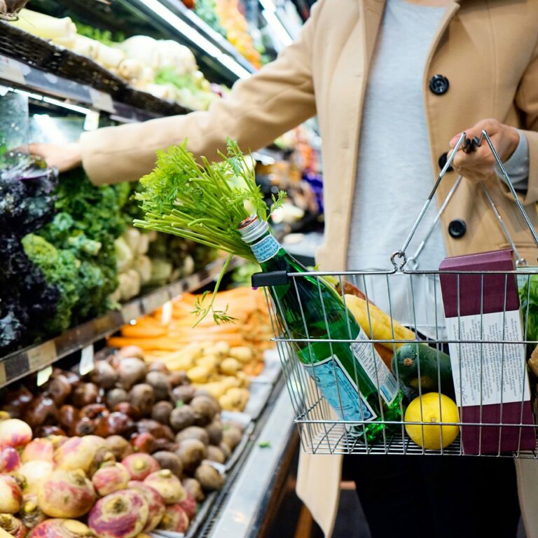 A shopping basket held by a person with vegetables and drinks in it